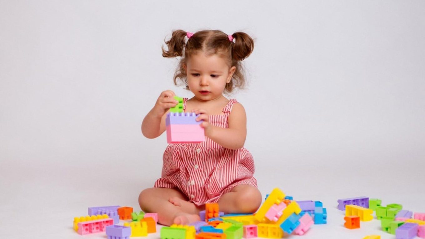A toddler girl sitting on the floor stacking colourful plastic building blocks
